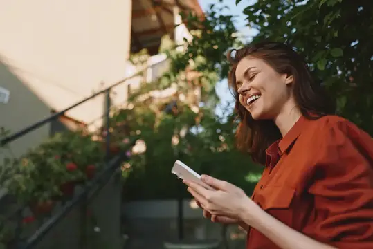 Улыбающаяся девушка в оранжевой рубашке смотрит в телефон на улице