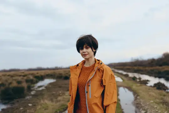Портрет женщины на отдыхе: оранжевая куртка, стоит на открытой тропинке через сельскую природу, ощущение прогулки, лицом к камере, деревенский сельский пейзаж, облачное пасмурное небо, мягкий свет
