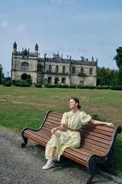 Молодая женщина в светлом платье отдыхает на скамейке перед старинным замком
