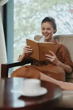 Улыбающаяся темноволосая женщина читает книгу в кресле у окна