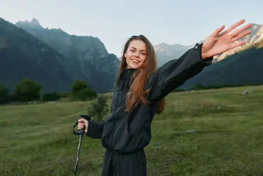 Улыбающаяся девушка с треккинговой палкой раскинула руки на фоне гор
