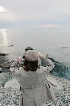 Вид со спины на женщину в пальто и берете на галечном пляже, обращенную к тихой воде