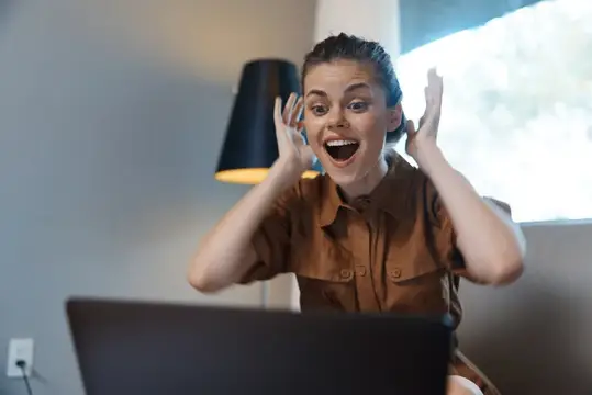 Радостная девушка с ноутбуком удивлённо вскинула руки дома
