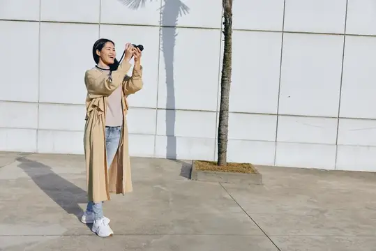 Улыбающаяся азиатская девушка фотографирует на камеру у белой стены