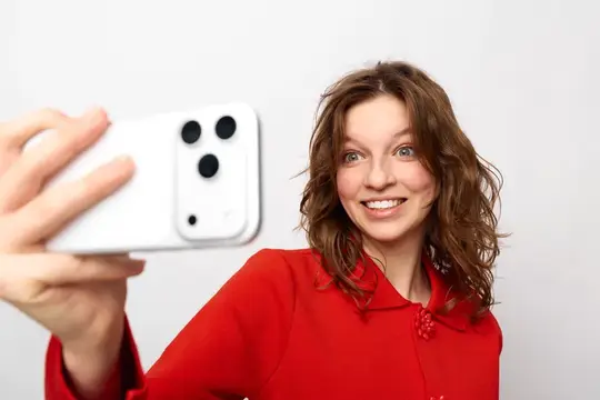 Счастливая молодая девушка в красном свитере держит белый смартфон, делая селфи на белом фоне
