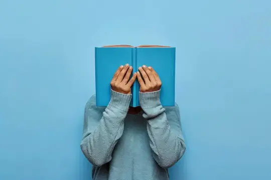 Человек в сером свитере закрывает лицо синей книгой на голубом фоне