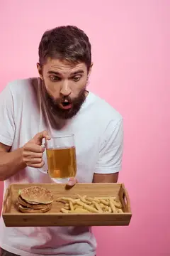 Удивлённый бородатый мужчина с пивом и подносом фастфуда на розовом фоне