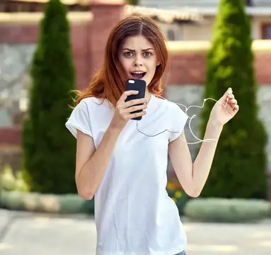 Рыжеволосая девушка в белой футболке удивлённо смотрит на телефон