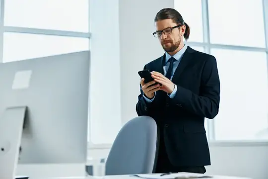 Бородатый мужчина в очках и деловом костюме смотрит в смартфон в офисе