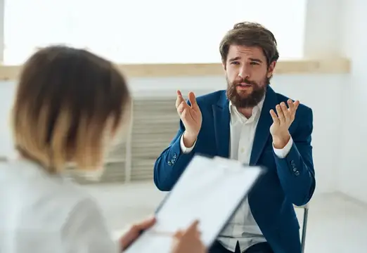 Бородатый мужчина в синем пиджаке эмоционально жестикулирует на встрече с коллегой