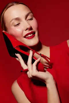 Улыбающаяся женщина с красной помадой и в красном платье держит красную туфельку на высоком каблуке близко к лицу