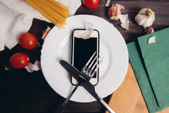 Смартфон на белой тарелке с приборами среди продуктов на тёмном столе