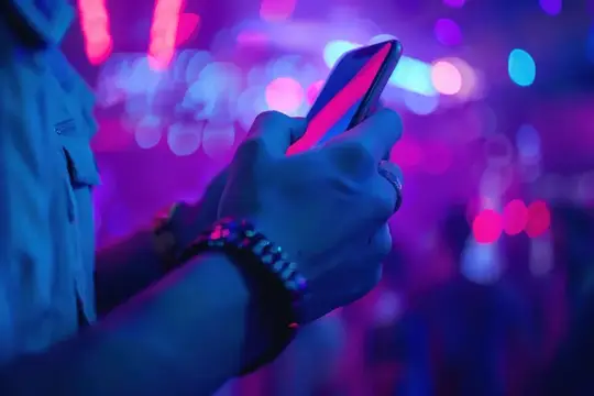 Руки человека с браслетом на запястье держат смартфон в неоновом свете ночного клуба
