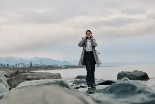 Молодая женщина в плаще стоит на камнях у моря в пасмурный день