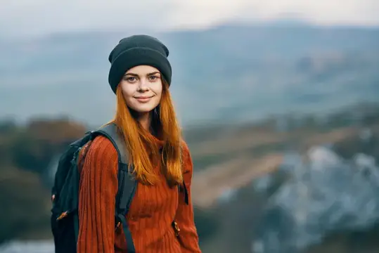 Рыжеволосая девушка в шапке с рюкзаком на фоне гор