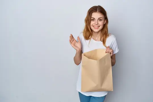 Улыбающаяся девушка с бумажным пакетом машет рукой на сером фоне