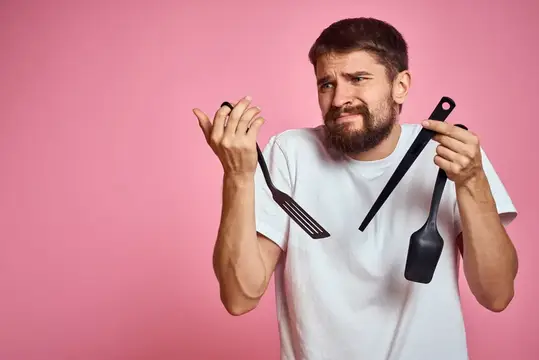 Бородатый мужчина с кухонными лопатками кривится на розовом фоне