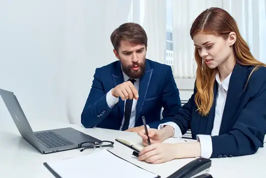 Мужчина и рыжеволосая женщина в деловых костюмах обсуждают работу за ноутбуком