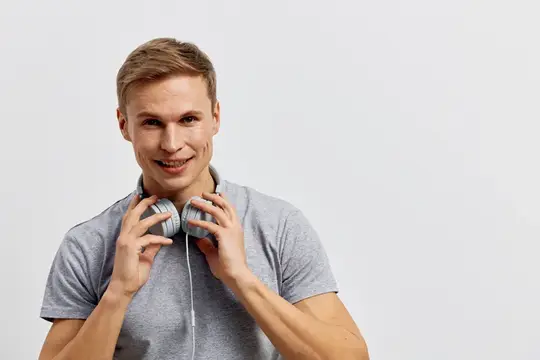 Улыбающийся светловолосый парень в серой футболке держит наушники