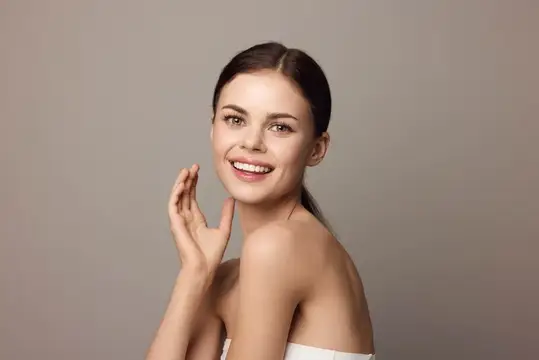 Улыбающаяся темноволосая женщина с ладонью у лица на сером фоне