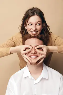 Улыбающийся мужчина с закрытыми глазами наслаждается игривым жестом женщины позади него, которая делает из рук очки
