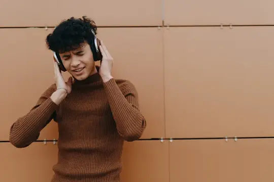 Модный молодой человек слушает музыку, улыбается, положив руки на голову, и стоит на фоне коричневой стены