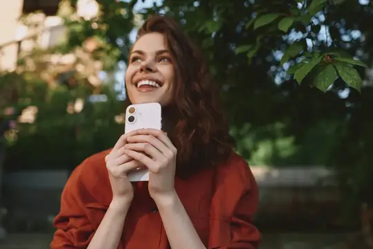 Непринужденная уличная сцена с улыбающейся женщиной и смартфоном, естественная парковая обстановка, расслабленная поза, повседневный момент использования технологий и счастья, яркая атмосфера