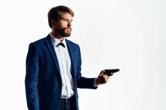 Бородатый мужчина в синем пиджаке держит пистолет на белом фоне