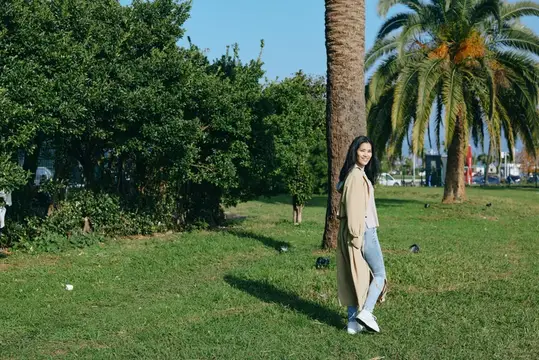 Молодая брюнетка в бежевом пальто позирует у пальмы в парке