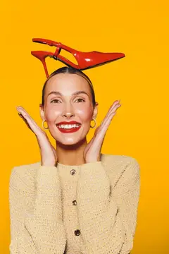 Счастливая женщина с красной туфлей на голове широко улыбается, на ней бежевый свитер и золотые серьги
