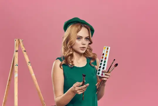 Молодая художница с палитрой и кистями у мольберта на розовом фоне