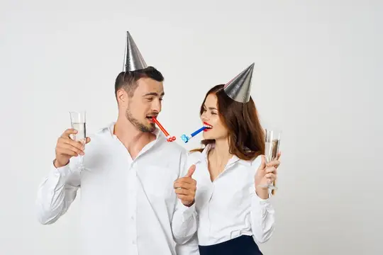 Мужчина и женщина в праздничных колпаках дуют в дудки с бокалами