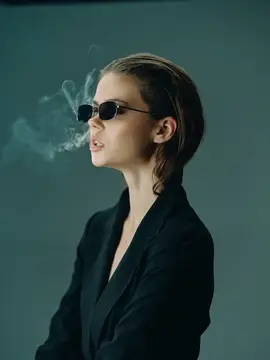 Молодая женщина в солнцезащитных очках выдыхает дым на тёмном фоне