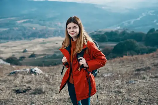 Улыбающаяся девушка с рюкзаком в красной куртке на горной тропе