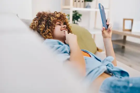 Кудрявая девушка лежит на диване со смартфоном и улыбается