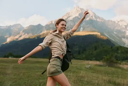 Улыбающаяся девушка с рюкзаком раскинула руки на фоне гор