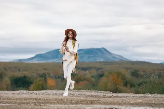 Улыбающаяся девушка в шляпе идёт по грунтовой дороге на фоне гор