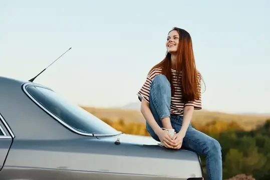 Женщина-водитель сидит на багажнике автомобиля и улыбается, любуясь красивым видом осенней природы и гор