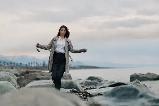 Молодая женщина в плаще идёт по камням у моря