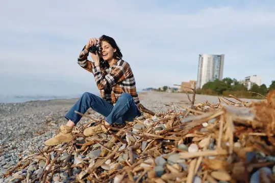 Улыбающаяся девушка с фотоаппаратом сидит на камнях у моря