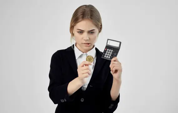 Светловолосая женщина в деловом костюме считает на калькуляторе монету