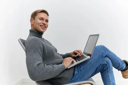 Улыбающийся мужчина в свитере работает за ноутбуком в кресле
