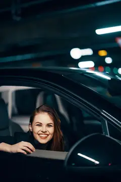 Улыбающаяся девушка выглядывает из окна автомобиля в тёмном паркинге