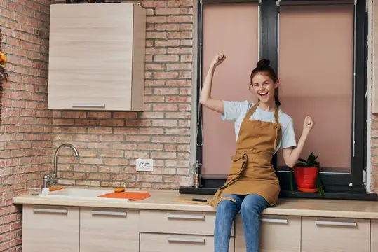Радостная девушка в фартуке сидит на кухонной столешнице с поднятыми руками
