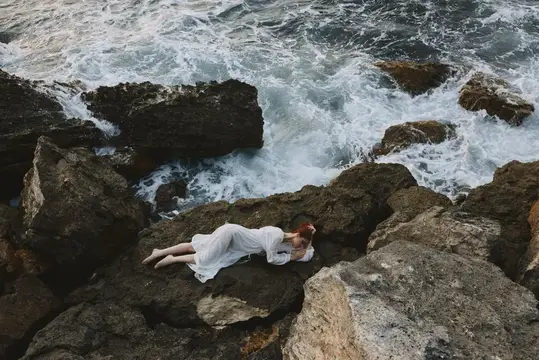 Женщина в белом платье лежит на камнях у бурного моря