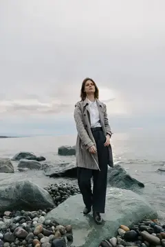 Молодая женщина в плаще стоит на камнях у пасмурного моря