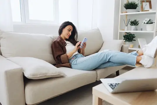 Брюнетка лежит на диване со смартфоном в руках