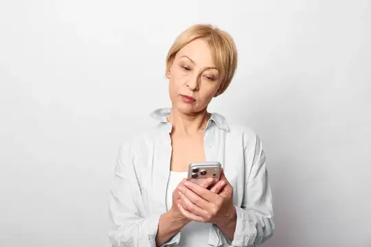 Блондинка в белой рубашке смотрит в смартфон на светлом фоне