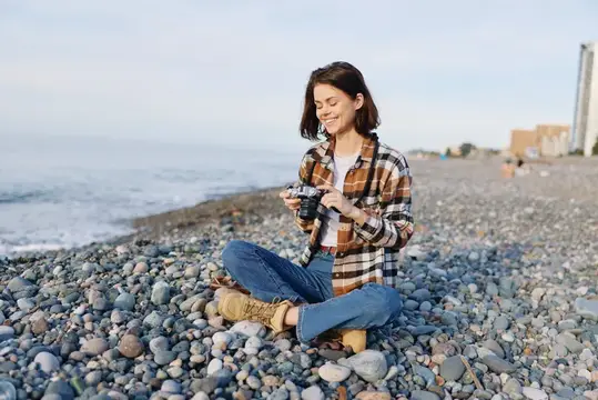Улыбающаяся девушка с пленочным фотоаппаратом сидит на галечном пляже