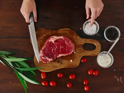 Женщина с ножом в руке нарезает свежий стейк для жарки на кухне, рядом соль, перец и другие специи на столе, красные помидоры черри и зелень, готовит ужин в домашней кухне